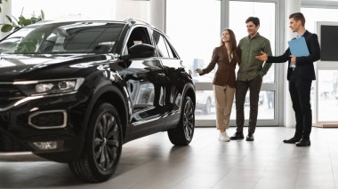 Confident car salesman showing new car to millennial couple at auto dealership, banner design