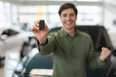 Handsome young guy showing new car key at camera, making YES gesture, celebrating purchase of auto at dealership