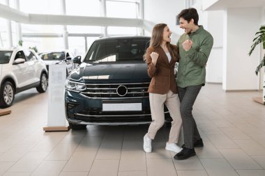Excited young couple making YES gesture, dancing and celebrating successful car purchase at auto dealership