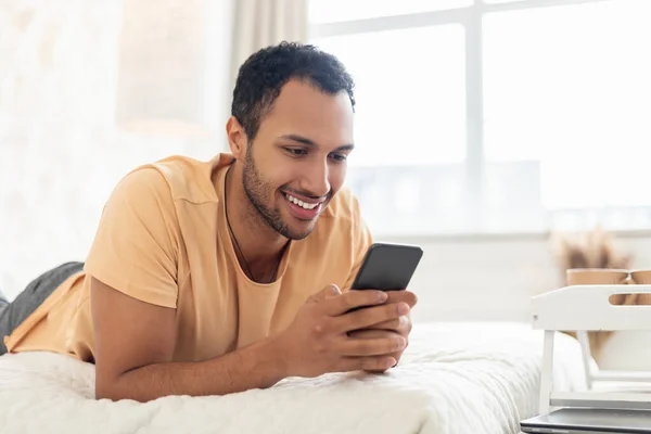 Happy Arabic Guy Using Phone Lying In Bed In Bedroom — стоковое фото