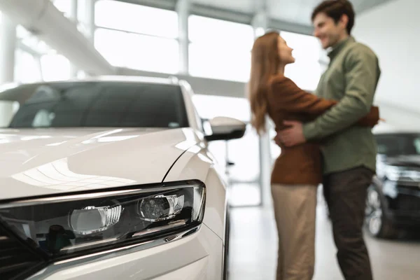 Happy young couple hugging at auto dealership, celebrating purchase of new vehicle, selective focus on car — Fotografia de Stock