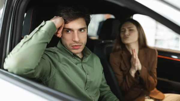 Unhappy young guy sitting inside car salon, his wife begging him to buy new auto at dealership, panorama — Stok fotoğraf