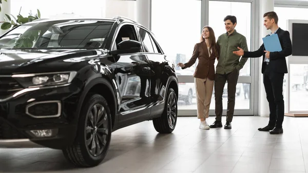 Confident car salesman showing new car to millennial couple at auto dealership, banner design