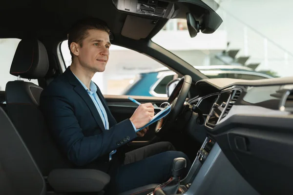 Confident young auto salesman sitting inside new car, making checkup, writing down information at dealership, copy space — 스톡 사진