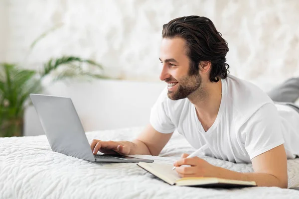 Positive young man laying on bed, using laptop, taking notes — Foto de Stock