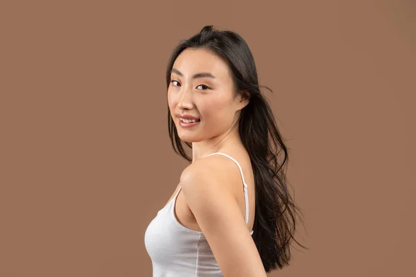 Schönheitsporträt einer hübschen asiatischen Dame mit glatter Haut und seidigen Haaren, die vor braunem Hintergrund posiert und in die Kamera blickt — Stockfoto