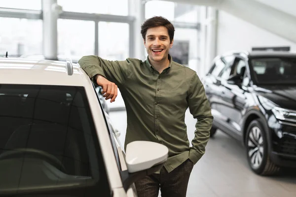 Jóképű, fiatal fickó pózol az új kocsija mellett, mosolyog a kamerába, autót vesz a kereskedőközpontban. — Stock Fotó