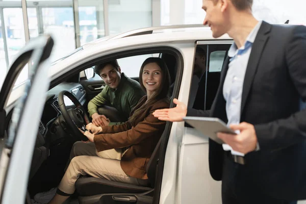 Millennial car salesman discussing purchase of new auto with happy young couple after test drive at dealership centre — ストック写真