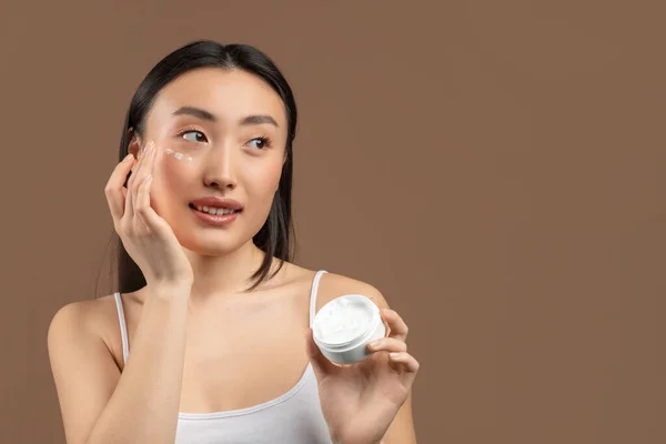 Portrait of young chinese lady with moisturizer cream on her pretty face, testing new beauty product, free space — Stockfoto