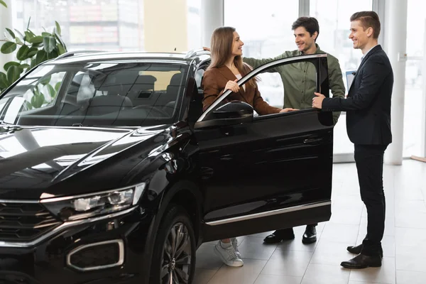 Cheery young couple speaking about buying new car with auto salesman at vehicle dealership — Stok fotoğraf