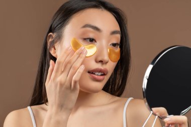 Genç Asyalı bayan ayna tutuyor ve göz bandı altında makyaj yapıyor, güzellik tedavisi yapıyor, kahverengi arka plan