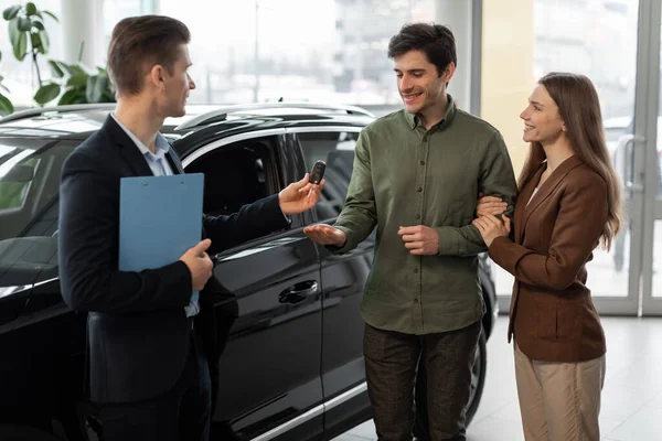 Car purchase or lease concept. Cheerful young couple taking car key from auto salesman at modern dealership — 스톡 사진