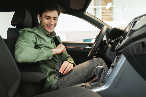 Young male client sitting at drivers seat of brand new car, fastening seatbelt, going on test drive at auto dealership — 스톡 사진