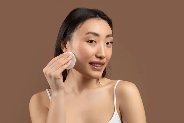 Sensuell asiatisk dam håller bomullspad för ansiktet, rengöring ansikte med toner över brun studio bakgrund — Stockfoto