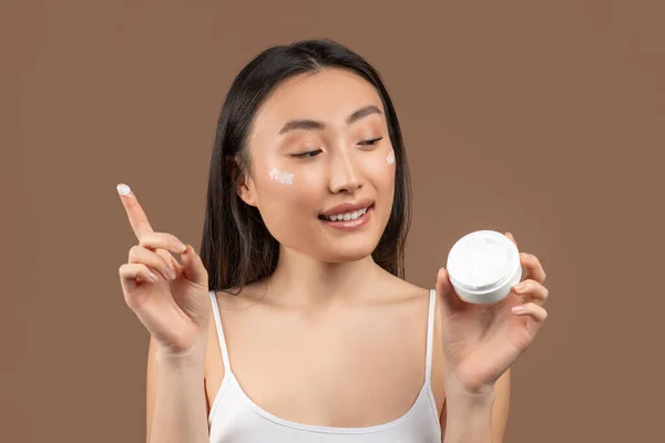 Skin care concept. Happy korean woman applying moisturizing cream on face, holding jar and smiling, brown background — Stockfoto