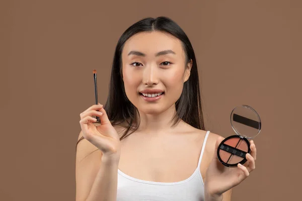 Everyday makeup. Portrait of pretty asian woman applying cosmetics, holding brush and palette, brown studio background — Stockfoto