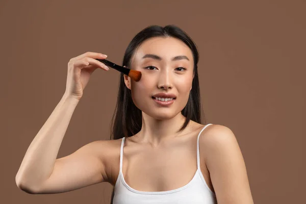 Attractive young asian lady with makeup brush applying blush on her cheeks, smiling at camera over brown background — Stockfoto