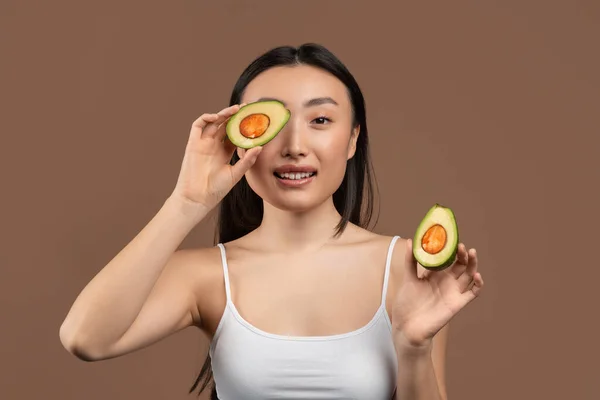 Beauty and anti-aging organic recipes concept. Pretty korean lady covering eye with half of avocado, brown background. — Zdjęcie stockowe