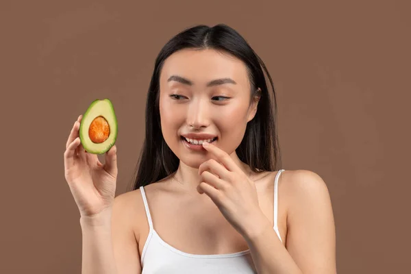 Natural skincare concept. Young asian woman holding ripe fresh avocado half, standing on brown background — Zdjęcie stockowe