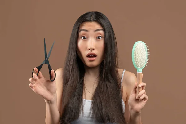 Shocked asian lady with long hair holding comb and metal scissors, thinking of professional treatment or fashion haircut — Photo