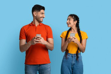 Zeki telefonlar tutan ve birbirlerine bakan neşeli genç bir Arap çift.