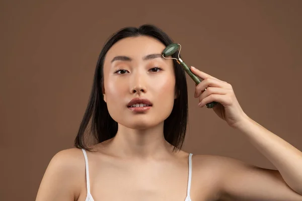 Beautiful asian lady using jade face roller on her flawless skin, looking at camera while posing over brown background — Stock Photo, Image