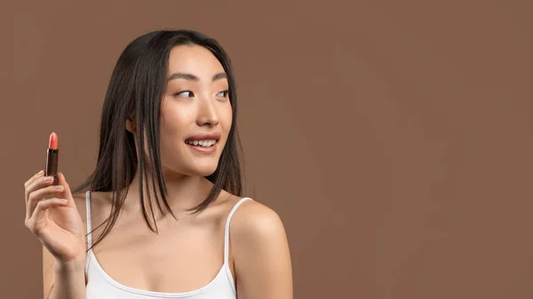 Attractive asian woman holding lipstick in hand, looking aside at free space and smiling over brown background, panorama — Stock Photo, Image
