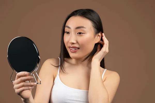 Conceito de cuidados com a pele. Bela mulher asiática olhando para o espelho e tocando seu cabelo sedoso, posando no fundo marrom — Fotografia de Stock
