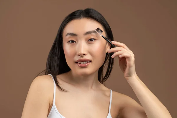 Charming asian lady applying eyebrow mascara, using decorative makeup and posing at camera over brown studio background — Stock Photo, Image