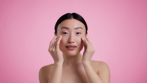 Cute young asian lady moisturizing her perfect skin, applying pampering serum, massaging her face, pink background — Vídeo de stock