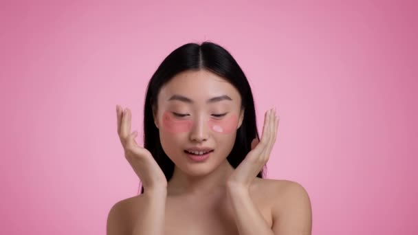 Procedimientos de belleza. Joven mujer asiática aplicando parches de colágeno bajo los ojos, mirando a la cámara, fondo rosa — Vídeos de Stock