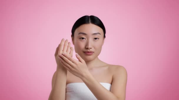 Protección de la piel y mimos. Joven hermosa mujer asiática aplicando crema hidratante en las manos, posando envuelta en toalla — Vídeos de Stock