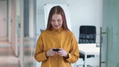 Young happy lady texting on smartphone and laughing, chatting with friends online, standing at modern office