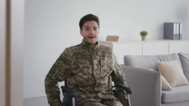 Kamuflajlı genç bir asker. Derin bir nefes alıyor ve kameraya gülümsüyor. Evde tekerlekli sandalyede oturuyor.