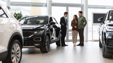 Car purchase or rental. Positive young spouses speaking to salesman about buying new auto at dealership, panorama