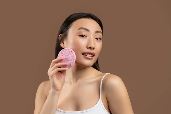 Skin care concept. Pretty asian lady holding cosmetic sponge, ready for face cleansing, posing over brown background — стоковое фото