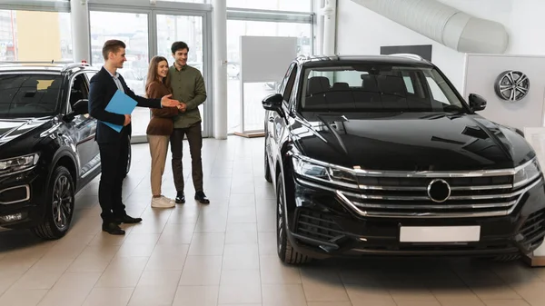 Confident car salesman showing modern luxury car to young Caucasian couple at dealership center, panorama