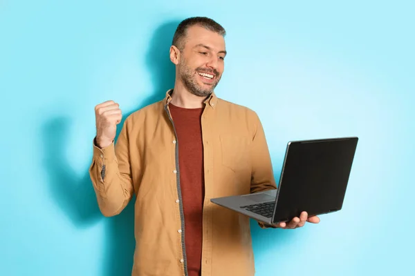 Homem alegre usando laptop balançando punhos em fundo azul — Fotografia de Stock