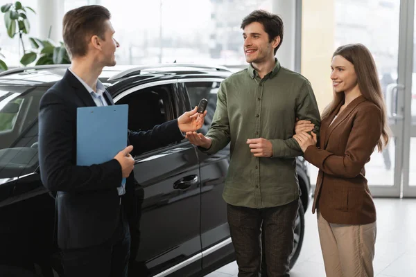 Positive auto showroom manager giving car key to happy young clients at car dealership — ストック写真