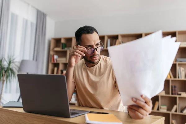 Serious young arab man reading documents and working on laptop remotely from home, sitting at desk — Foto de Stock