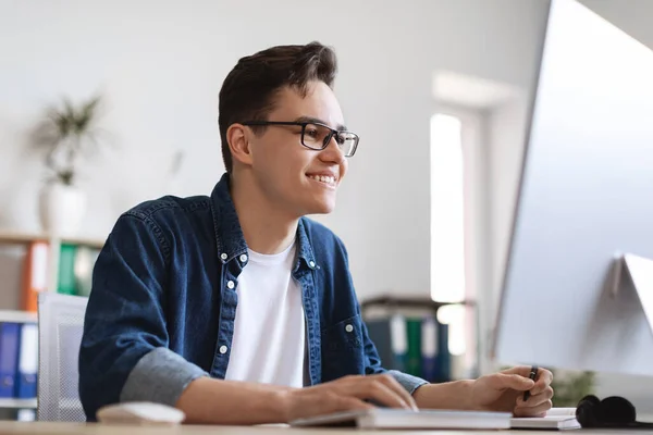 Junge männliche Büroangestellte machen sich Notizen, während sie am Schreibtisch am Computer arbeiten — Stockfoto