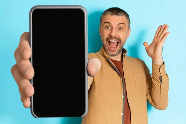 Aufgeregter Mann zeigt leeren Handybildschirm und schreit über blauen Hintergrund — Stockfoto