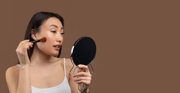 Pretty asian woman applying blush on her cheeks with makeup brush, looking at mirror, brown background, panorama — Stockfoto