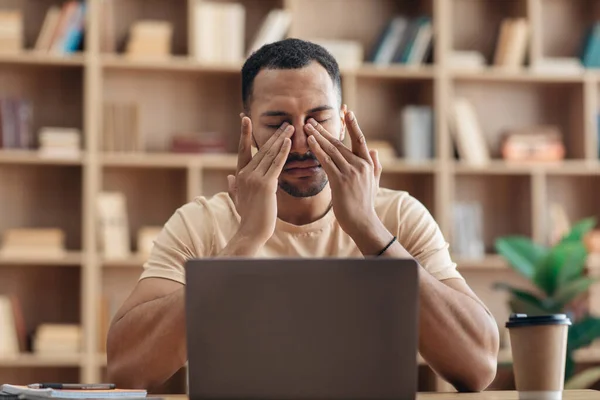 Tired arab freelancer guy sitting at workdesk in front of laptop at home and rubbing his eyes, suffering from burnout — Stockfoto
