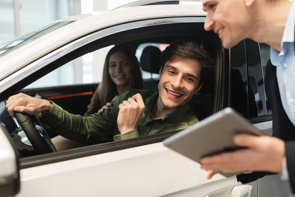 Young Caucasian couple clarifying car purchase details with salesman, looking at tablet screen in dealership center — 스톡 사진