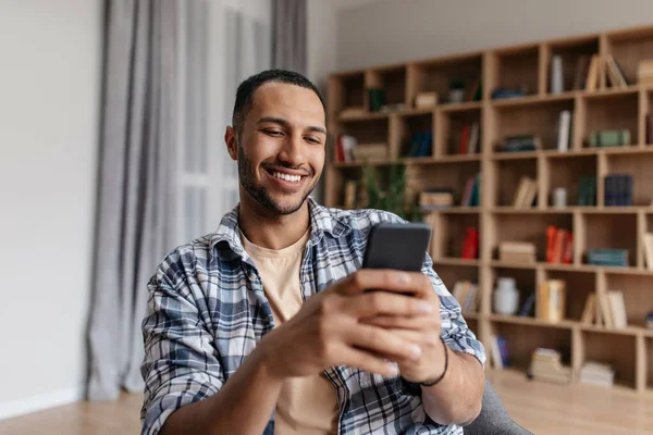 Užívám si volného času. Šťastný arabský muž pomocí smartphone zatímco sedí na židli doma, relaxaci v obývacím pokoji — Stock fotografie