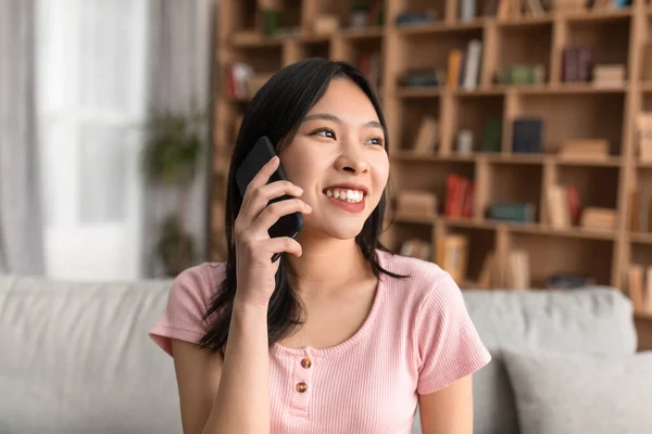 Alegre dama coreana hablando en el teléfono inteligente mientras está sentado en el sofá en casa, mirando a un lado y sonriendo, espacio de copia — Foto de Stock