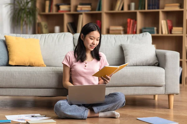 Young korean lady studying online from home, reading book and using laptop pc, sitting on floor near sofa, free space — Foto de Stock