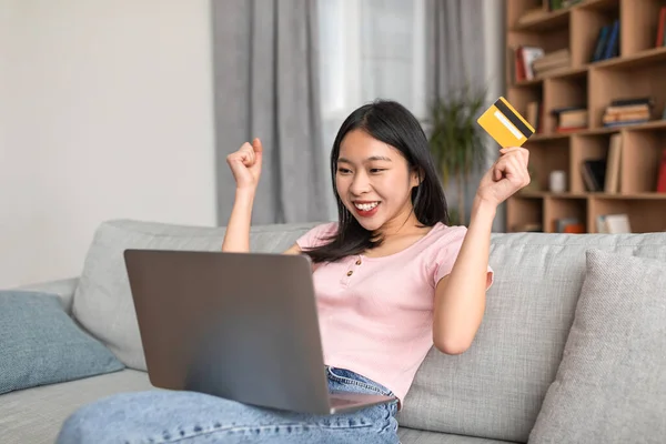 Nadšený korejská dáma s notebookem a kreditní kartou gestikulující ANO, šťastný nad obrovskou slevu, odpočívá na pohovce doma — Stock fotografie