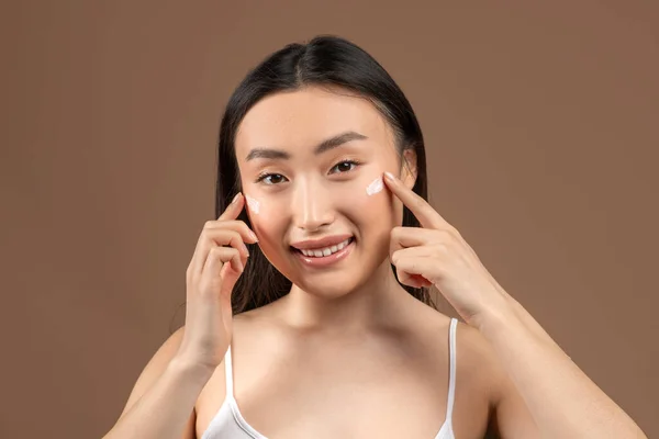 Attraktive junge asiatische Dame berührt ihre glatten Wangen, trägt Feuchtigkeitscreme auf und lächelt in die Kamera — Stockfoto